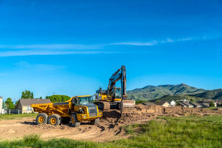 Truck and excavator at a construction site with blue sky overhead on a sunny day. Houses and distant mountain can be seen in the background.の写真素材