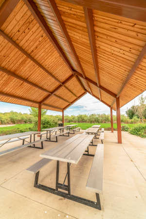 Row of white tables with seats under a pavilion with brown ceiling at a park. The picnic area has a scenic view of vibrant foliage and cloudy blue sky.の写真素材