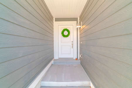 Narrow corridor with gray wood wall leading to the white front door with wreath. A bright wall lamp...