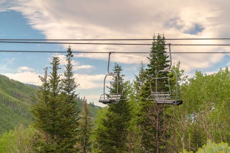 Chairlifts and green trees against cloudy blue sky in Park City Utah in summer. The mountain in this ski resort is covered with lush foliage during off season.の写真素材