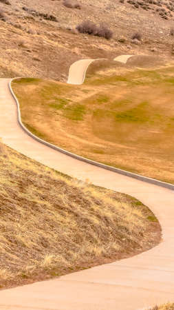 Vertical frame Winding narrow rural road through rolling foothills of snow covered mountains surrounded by fields with dry grass after winter in a scenic landscapeの写真素材