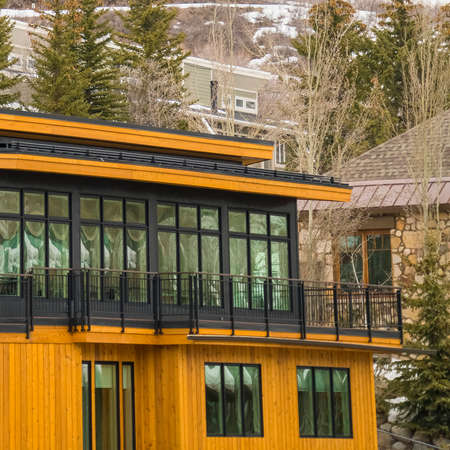 Square Modern home exterior with yellow wall balcony flat roof and large windows. Coniferous trees,...