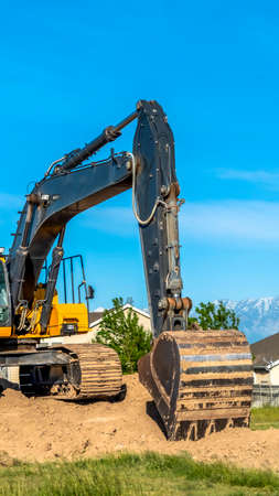 Vertical Close up of an excavator with arm bucket and continuous tracks on top of soil. Houses and blue sky can be seen from the construction site.の写真素材
