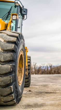 Vertical Close up of the tires of a bulldozer against snow peaked mountain and cloudy sky. The construction vehicle has yellow paint, empty cab, and front lights.の写真素材