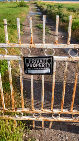 Vertical Locked rusty old gate with private property sign and view of lake and mountain. Dirt road with puddles of water amid grassy terrain can be seen behind the gate.の写真素材