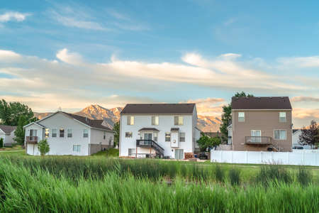 Pond surrounded by green grasses with homes and mountain against cloudy blue sky. Nature and...