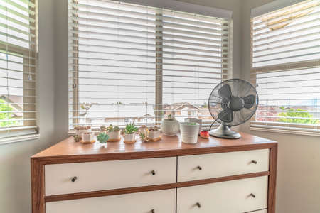 Home interior with potted cacti and electric fan on cabinet against bay window. Houses and sunny outdoors can be seen through the white window blinds.の写真素材