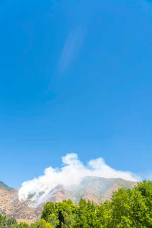 Scenic nature view on a sunny day with smoke from fire in the mountains. Wild forest fire with smoke rising into the clear blue sky.の写真素材