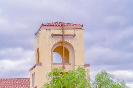 Modern tiled bell tower of a small church decorated with a simple cross rising above green trees