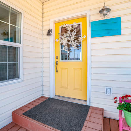 Square Bright yellow front door to a white wooden house decorated with a spray of flowers under a covered porchの写真素材