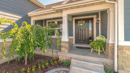 Pano frame Home with landscaped yard stairs porch and front door with sidelight and wreath. Window, chair, gray wall siding, and stone brick wall can also be seen at the facade of this house.