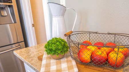 Pano Fruit basket with red apples on top of chopping board and marble countertop. Kitchen interior of a home with white cabinets, gray wall, and refrigerator.の写真素材