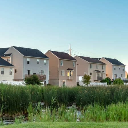 Square Green grasses and pond at a scenic park with houses and blue sky background. Balconies, stairs, fences, and lawns can be seen at the facade of the homes.の写真素材