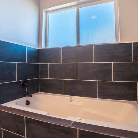 Square Modern bathroom with dark grey tiles surrounding a fitted tub below a small windowの写真素材