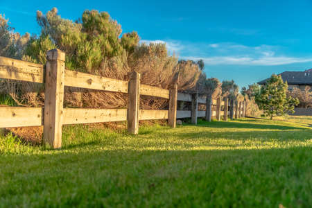Brown wooden low fence on a lush green field against thick bushes and shrubs. Sunny day view with...