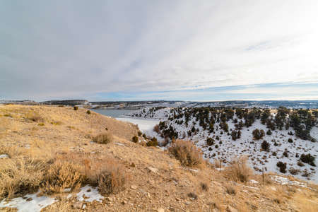 Scenic winter landscape with calm lake amid hills blanketed with white snow. Vast blue sky filled...