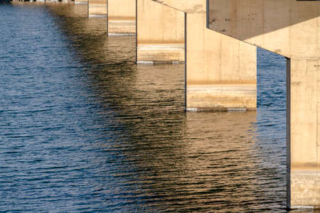 Views underneath a beam bridge with abutments against the sunlit lake water. The massive piers of a...
