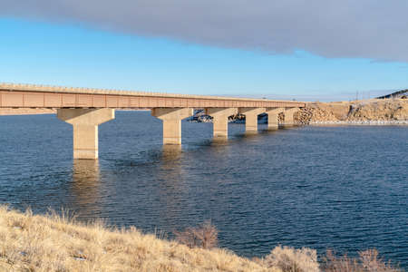 Bridge across blue lake with views of snow covered hills and cloudy blue sky. A snowy sloping...