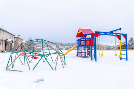 Snow covered park with playground against homes hills and cloudy sky in winter. The colorful slide,...