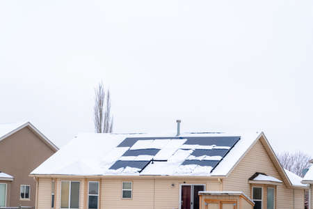Snow covered roof of home with solar panels against cloudy sky in winter. Bay window and white...