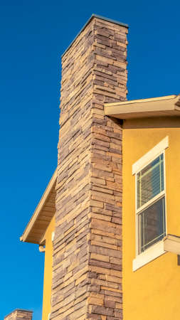 Vertical Home exterior with stone brick chimney against gable roof and yellow wall. A view of vibrant blue sky and treetop can be seen in the background on this sunny day.の写真素材
