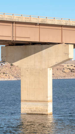 Vertical frame Bridge and pier foundation over blue lake water against mountain and blue sky. A...