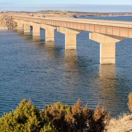 Square frame Blue lake with bridge connecting vast terrains blanketed with snow in winter. Green...
