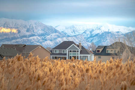 Homes and stunning snowy mountain with blurry brown grasses in the foreground. A ray of sunlight beams through the cloudy sky and illuminates the mountain slope.の写真素材