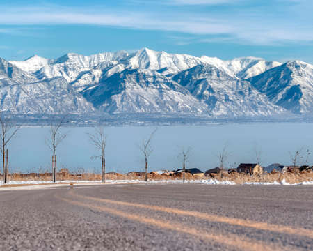 Road lined with young trees overlooking snowy Wasatch Mountains and Utah Lake. Houses on the...
