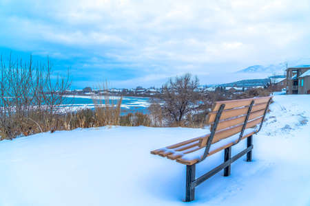 Empty outdoor bench on snowy terrain overlooking frozen Utah Lake in winter. Houses on a lake side neighborhood and distant mountain can also be seen in this landscape.の写真素材