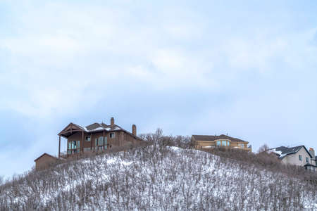 Homes on the peak of a hill with brown leafless bushes on snowy slope in winter. The neighborhood has scenic views of frosted nature and overcast sky.の写真素材