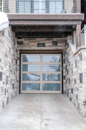 Snow falling down on home with glass panel garage door and stone exterior wall. Balcony and stairs...