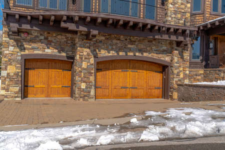 facade of home featuring two hinged wooden garage doors and stone brick wall. A balcony with metal railing has a view of the snowy road on a sunny winter day.の写真素材