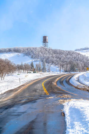 Paved road on the snowy slopes of Wasatch Mountains with cloudy blue sky view. Frosted trees, evergreens, and water tower can also be seen in this winter landscape.の写真素材