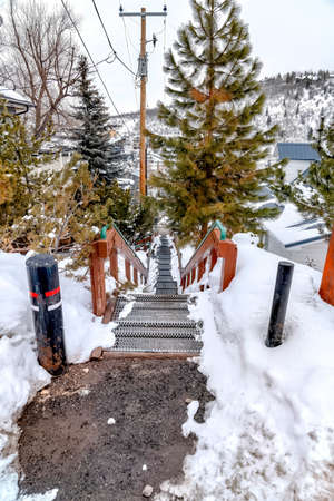 Stairs overlooking residential homes and scenic hill blanketed with winter snow. Grate metal stairs with handrails surrounded with trees and electric posts.の写真素材
