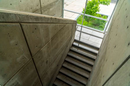 Commercial building interior with staircase and handrails between concrete walls. The clear glass wall on the landing has a view of a vibrant young tree.の写真素材