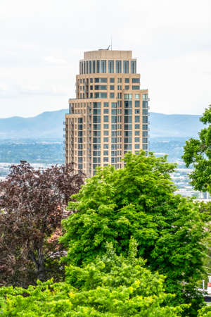 Lush natural tree foliage against towering modern skyscraper building. The valley, mountain, and cloudy sky can be seen in the blurry background.の写真素材