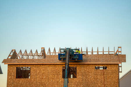 Builders roofing a new build house using a mechanical arm with exposed trusses against a blue skyの写真素材
