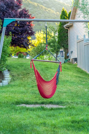 Backyard of home with red hammock hanging on metal pole and lush green grasses. Abundant foliage and blurry hill can be seen in the background.の写真素材
