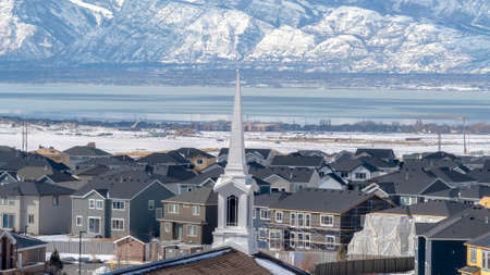 Panorama Church spire against neighborhood homes with snowy mountain and scenic lake view. Blue sky and puffy clouds can be seen over the residential landscape blanketed with snow in winter.の写真素材