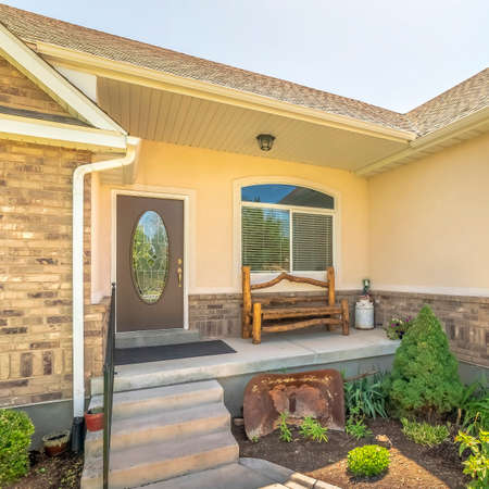 Square crop Home facade with view of arched garage door and front door with oval glass pane. A pathway connects the driveway to the steps that leads to the porch with furniture against window.の写真素材