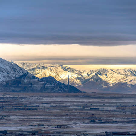 Square frame Sweeping view of Salt Lake City bordered by towering sunlit snowy mountain. A secnic panoramic Utah landscape with cloudy sky overhead.の写真素材