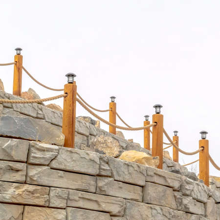 Square frame Rope fence with lamps on posts lining a retaining wall made of stone blocks
