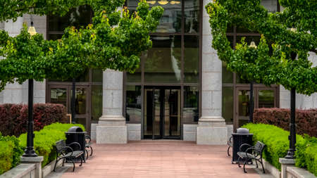 Panorama crop Entrance to a white building with pavement leading to the glass door entryway. Trees, shrubs, benches, and garbage bins can be seen on the sides of the walkway.の写真素材