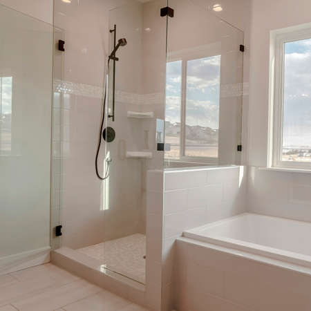 Square Built in bathtub with black faucet and shower stall with half glass enclosure. Interior of bathroom with view of snowy landscape and cloudy sky through the window.の写真素材