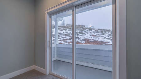 Panorama View through glass doors leading to a small exterior balcony of winter snow in a rural landscape with distant houseの写真素材