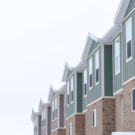 Square crop Upper storey of townhomes with brick wall vertical siding and snowy roofs. Residential building exterior viewed against cloudy sky on a cold winter day.の写真素材