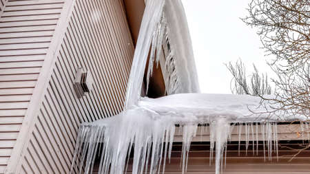 Panorama Roof of home with thick snow and sharp icicles against cloudy winter sky. The wall of the...