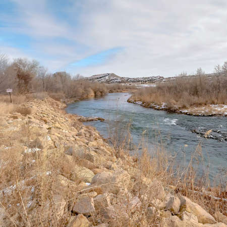Square frame Scenic river with snowy shore against distant hills under cloudy sky in winter. Rocks,...