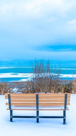 Vertical crop Bench overlooking semi frozen Utah Lake with overcast sky over the horizon. The empty outdoor seat in on a hilly terrain covered with snow in winter.の写真素材